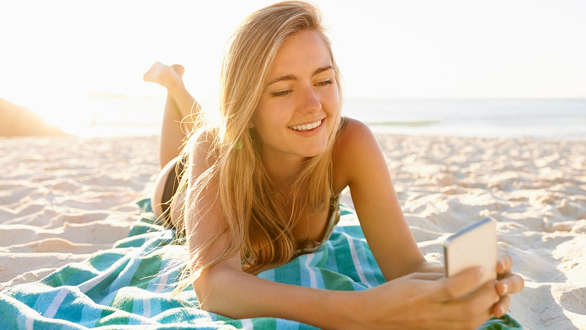 woman using her cellphone while lying on a beach