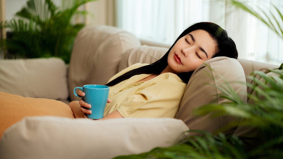 A relaxed woman takes a nap on a couch, holding a mug of coffee